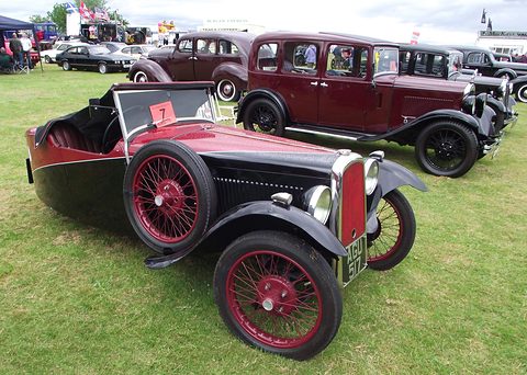 A black and red three-wheeler with red wire wheels on grass in front of a row of vintage saloons. Opens a larger view