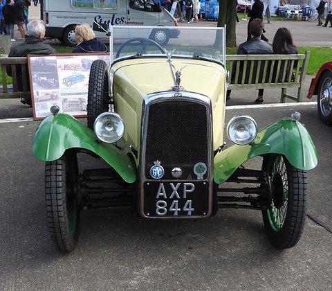 Front view of a cream three-wheeler with green wings, a black mesh radiator and club badges. Opens a larger view