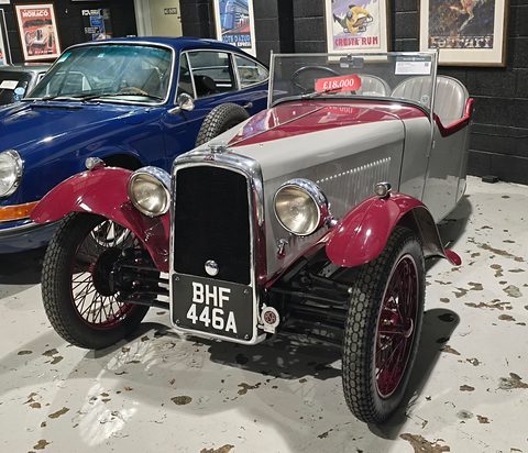 A grey three-wheeler with maroon wings, wire wheels and a chrome radiator on a wet floor, a blue Porsche behind. Opens a larger view