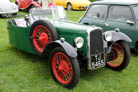 A green three-wheeler with red wire wheels and a chrome radiator on grass beside a Mini. Opens a larger view