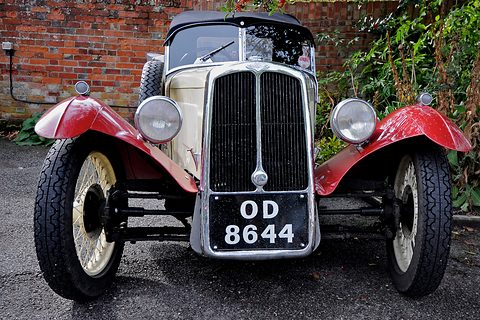Front view of a cream three-wheeler with red wings, a tall chrome radiator and cream wire wheels before a brick wall. Opens a larger view