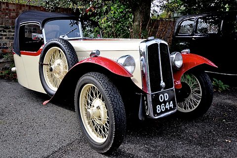 A cream and red three-wheeler with a black hood, wire wheels and a spare wheel at its side on tarmac. Opens a larger view