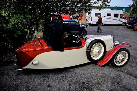 Side-rear view of a cream and red three-wheeler with a pointed red tail, a black hood and a spare wheel. Opens a larger view