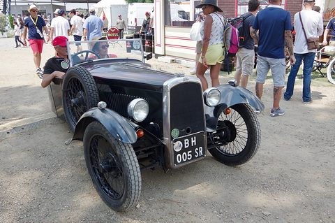 A black open three-wheeler with wire wheels and a chrome radiator, a driver aboard, among visitors in a paddock. Opens a larger view