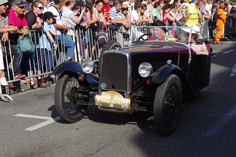 A black three-wheeler with a parade plate on its front driving past a crowd behind barriers. Opens a larger view