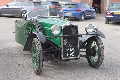 Front view of a green three-wheeler with a black mesh radiator, two headlamps and green wire wheels. Opens a larger view