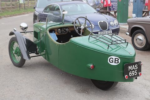 Rear view of a green three-wheeler with a luggage rack and a GB plate on its rounded tail, parked cars behind. Opens a larger view