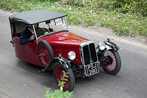 A red three-wheeler with a black hood and a driver aboard climbing a narrow lane between grass verges. Opens a larger view