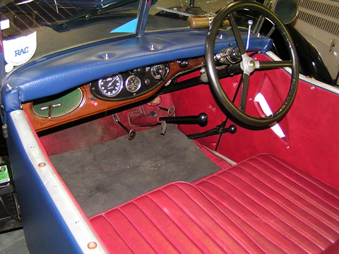 A four-spoke steering wheel before a wooden dash with round gauges, above a red leather bench seat. Opens a larger view