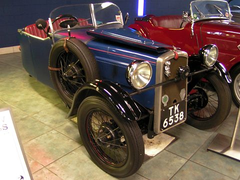 A blue three-wheeler with black wings, a chrome radiator and two headlamps beside a red sports car. Opens a larger view