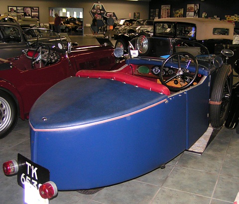 Rear three-quarter view of a blue three-wheeler with a pointed tail, red seat and rear lamps beside the plate. Opens a larger view