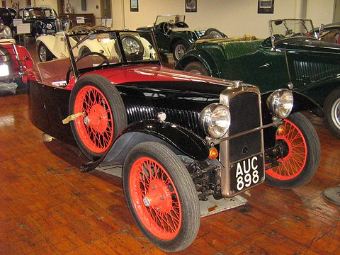 A black three-wheeler with a red cockpit, red wire wheels and plate AUC 898 on a wooden floor. Opens a larger view
