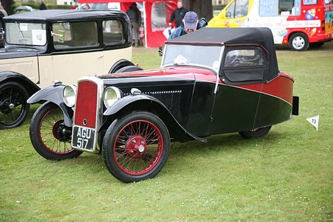 A black and red three-wheeler with a raised black hood and red wire wheels on grass, show tents behind. Opens a larger view