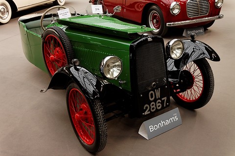 A green three-wheeler with black wings, red wire wheels and a black radiator, a Bonhams sign on the floor before it. Opens a larger view