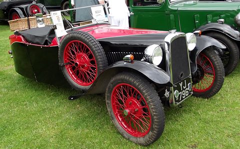 A black three-wheeler with red panels and red wire wheels on grass, a picnic basket on its tail. Opens a larger view