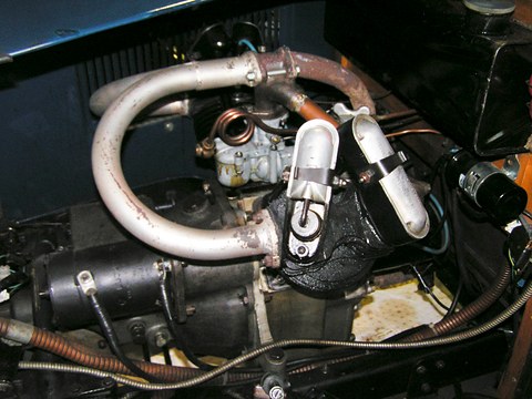A V-twin engine with a looping exhaust pipe, carburettor and finned cylinders under a lifted bonnet. Opens a larger view