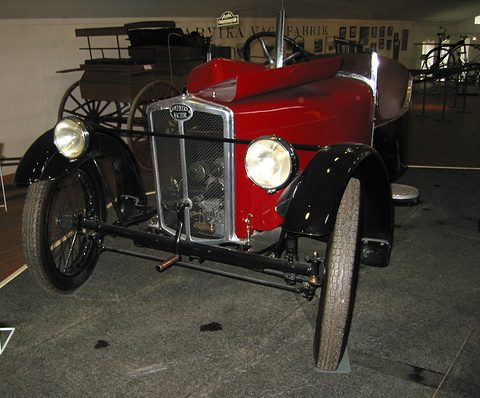 Front view of a red three-wheeler with a chrome mesh radiator, two headlamps and two front wheels on a dark floor. Opens a larger view