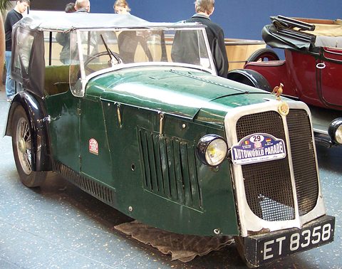 A green three-wheeled open car with a fabric hood, a chrome radiator and a rally plate, on a show floor. Opens a larger view