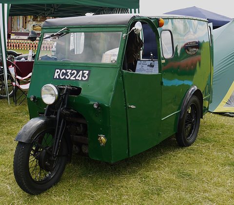 A green van body on two rear wheels with a motorcycle-style front fork and wheel, on grass by a marquee.