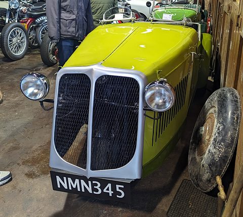 Front view of a yellow-green three-wheeler with a split chrome radiator and plate NMN 345 in a shed.