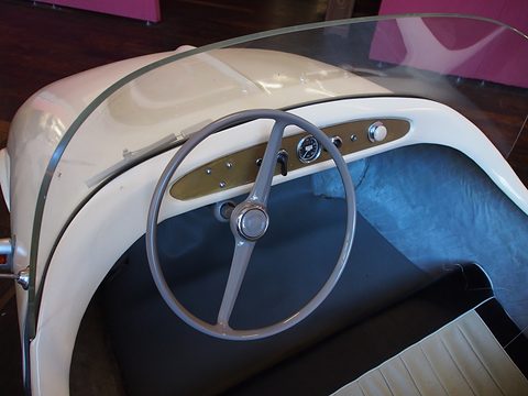 Thin two-spoke steering wheel in front of a cream dashboard with small round instruments, behind a curved screen. Opens a larger view