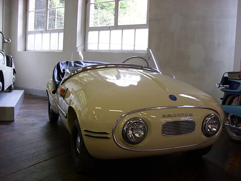 Cream open roadster with BRÜTSCH lettering above its grille, on a dark wooden floor by tall windows. Opens a larger view