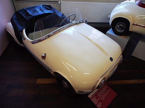 Small cream open two-seater with hood stowed under a black tonneau, on a museum floor.
