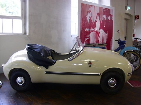 Cream open two-seater side-on, hood folded behind the seats, a red poster on the wall behind. Opens a larger view
