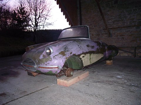 Flaking purple and green open sports body without wheels, resting on wooden blocks under a barn roof.