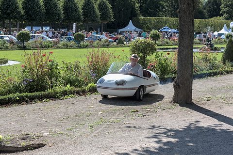 A small white egg-shaped car with a driver in a white hat on a gravel path beside flower beds. Opens a larger view