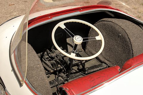 White three-spoke steering wheel above the pedals, a red seat and carpeted tub. Opens a larger view