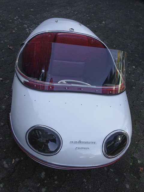 White rounded nose with two oval lamp housings and a curved clear screen, on dark tarmac. Opens a larger view