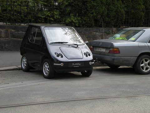 Black rounded microcar with four small round headlamps parked at a kerb beside a silver Mercedes saloon.