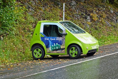 Side view of a lime-green rounded microcar on a wet road below a grassy, rocky bank. Opens a larger view