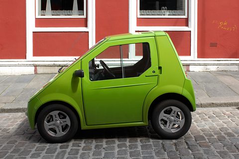 Side view of a lime-green rounded microcar on cobbles in front of a red-painted wall and white windows. Opens a larger view