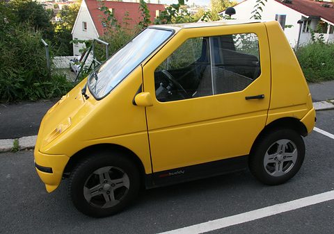 Side view of a yellow rounded three-seat microcar with alloy wheels in a marked bay beside shrubs. Opens a larger view