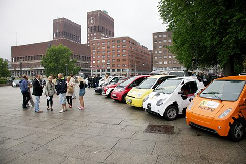 An orange Buddy at the head of a line of small electric cars on a paved square, City Hall towers behind. Opens a larger view