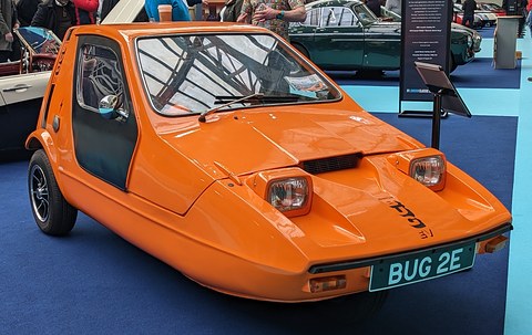 Tangerine three-wheeler on blue carpet at an indoor show, visitors at the edge of the frame. Opens a larger view