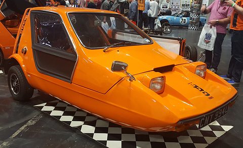 Tangerine three-wheeler on a chequered floor in a busy show hall.