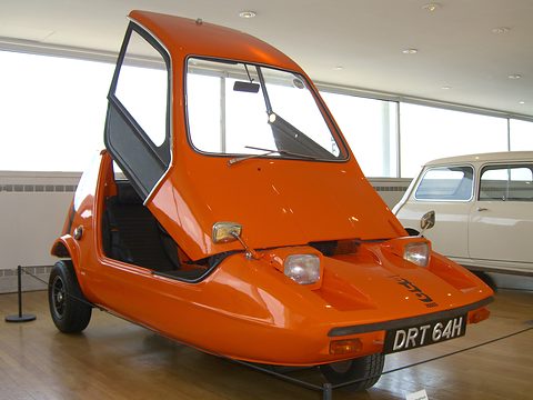 Tangerine three-wheeler with its canopy up on a wooden museum floor, a cream classic car behind. Opens a larger view