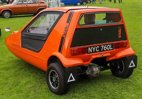 Tangerine three-wheeler from behind, black tail panel with Bug script and the rear axle visible on grass. Opens a larger view
