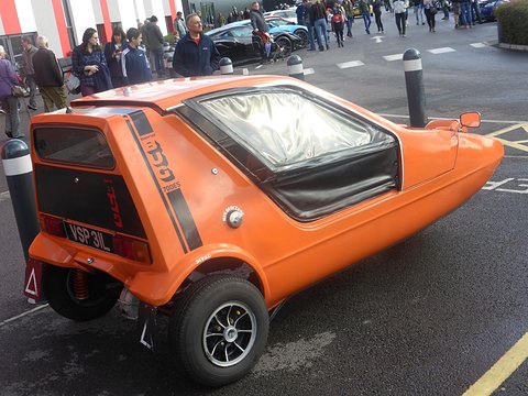 Tangerine three-wheeler from the rear quarter on a car park, visitors walking behind. Opens a larger view
