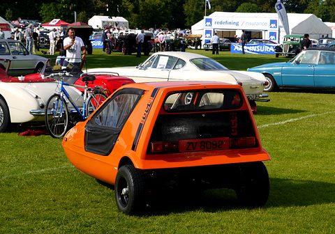 Tangerine three-wheeler from the rear quarter on a show lawn, marquees and visitors behind. Opens a larger view