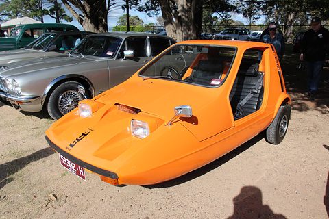 Tangerine wedge three-wheeler on dusty gravel in sunlight, a silver saloon behind it. Opens a larger view