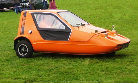 Tangerine three-wheeler side-on on a green field with its driver inside, spectators behind a tape. Opens a larger view