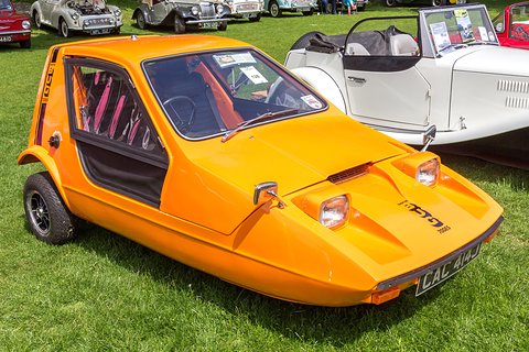 Tangerine three-wheeler on grass beside a white open roadster. Opens a larger view