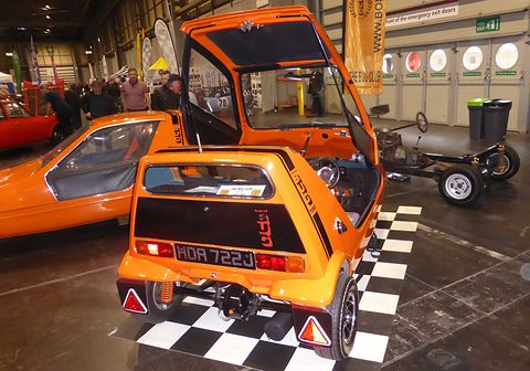Tangerine three-wheeler seen from behind with its canopy lifted, a bare chassis on the chequered floor beyond. Opens a larger view