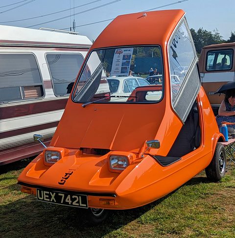 Tangerine three-wheeler seen head-on with its roof and windscreen lifted up as one piece. Opens a larger view