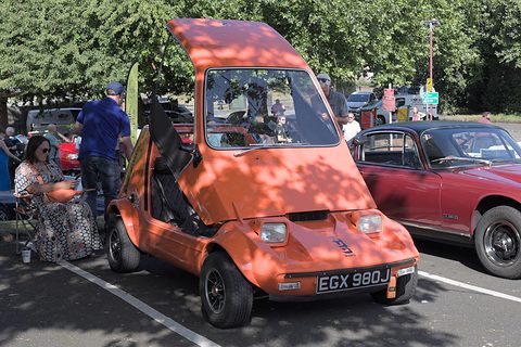 Tangerine three-wheeler in dappled shade with its canopy raised, a maroon car and visitors beside it. Opens a larger view