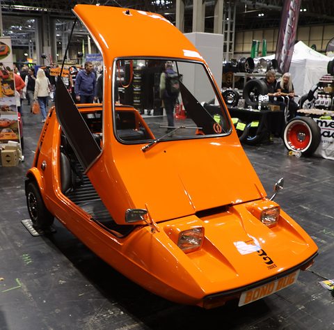 Tangerine three-wheeler head-on with its canopy raised in a crowded exhibition hall. Opens a larger view